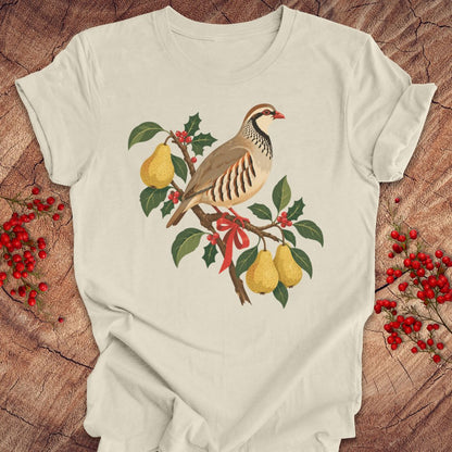 Natural cream t-shirt with a partridge perched on a branch with golden pears, holly, red berries, and a red ribbon, laid flat on a rustic wood background with red winter berries.