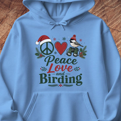 A light blue hoodie laid flat on a rustic wood background, featuring a festive front graphic with a peace sign, red heart, bird, binoculars, pine branches, and the words 'Peace Love and Birding' in mixed dark and red lettering.