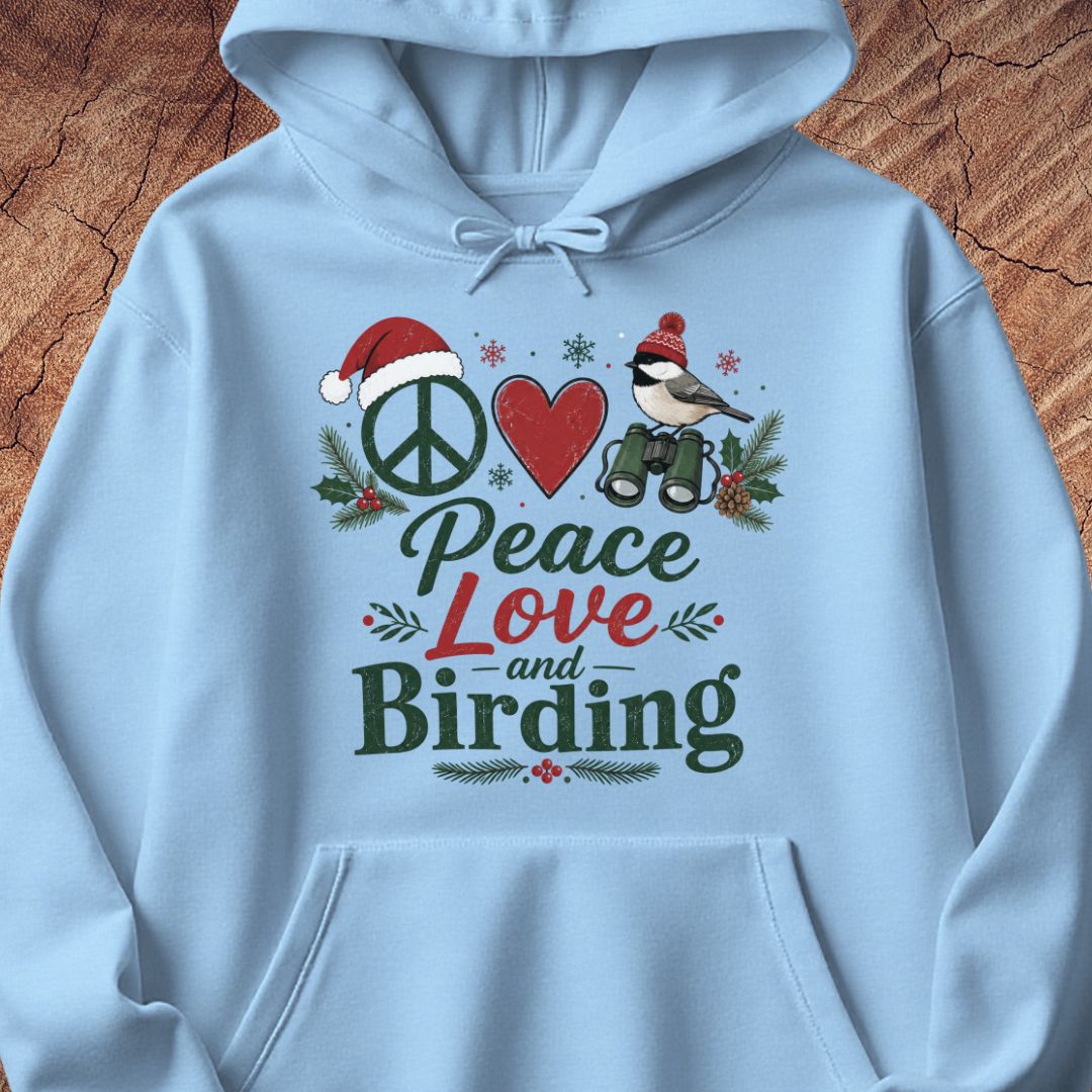 A light blue hoodie laid flat on a rustic wood background, featuring a festive graphic print with a peace sign, red heart, bird, binoculars, pine branches, and the words 'Peace Love and Birding'.