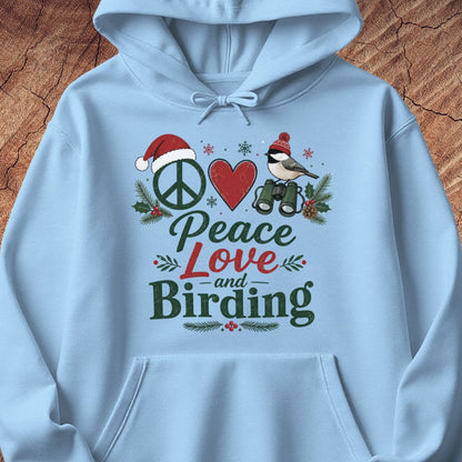 A light blue hoodie laid flat on a rustic wood background, featuring a festive graphic print with a peace sign, red heart, bird, binoculars, pine branches, and the words 'Peace Love and Birding'.