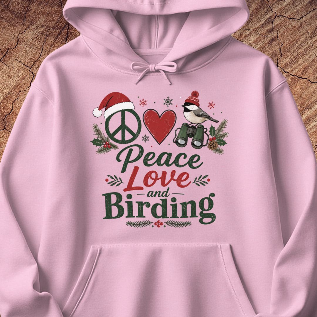 A light pink pullover hoodie with a festive graphic print reading 'Peace Love and Birding' in dark green and red lettering, decorated with a peace sign, red heart, bird, Santa hat, and holiday holly, shown on a wood background.