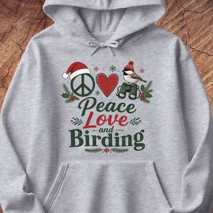 A light gray heather pullover hoodie with a festive Christmas-themed graphic on the chest. The graphic features a peace sign wearing a Santa hat, a red heart, a bird with binoculars, holly leaves, and the text 'Peace Love and Birding' in green and red lettering.
