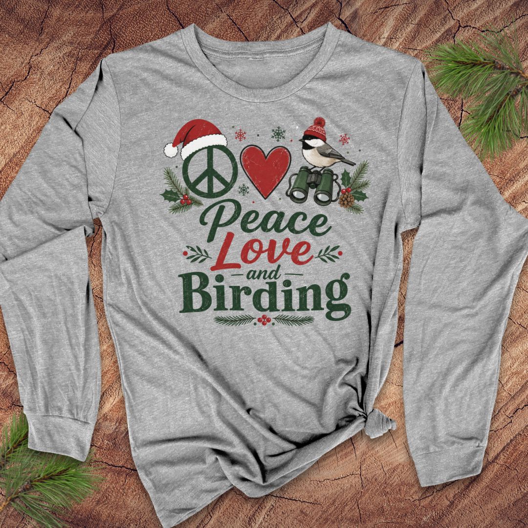 A heather gray long-sleeve t-shirt with a festive graphic print reading 'Peace Love and Birding' in green and red lettering, featuring a peace sign, red heart, bird, and holiday pine accents, laid flat on a rustic wood background with pine needles.