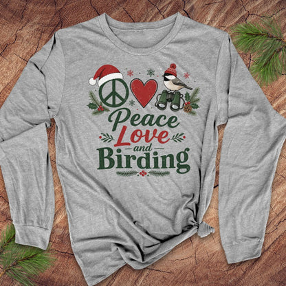 A heather gray long-sleeve t-shirt with a festive graphic print reading 'Peace Love and Birding' in green and red lettering, featuring a peace sign, red heart, bird, and holiday pine accents, laid flat on a rustic wood background with pine needles.
