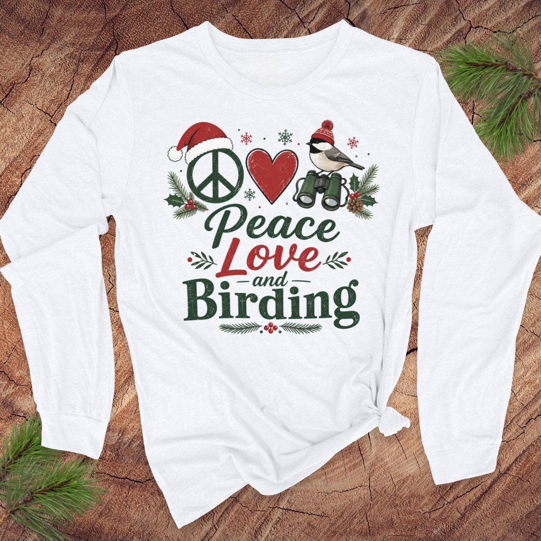 A white long-sleeve t-shirt with a Christmas-themed graphic print reading 'Peace Love and Birding' in green and red lettering, surrounded by a peace sign, heart, bird, and pine branches, laid flat on a rustic wood background with pine needles.
