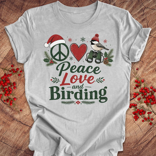 A light gray short-sleeve t-shirt with a festive Christmas-themed graphic that reads 'Peace Love and Birding'. The graphic features a peace sign with a Santa hat, a red heart, a bird with binoculars, and holiday greenery.