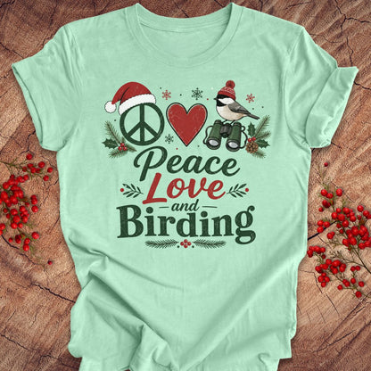 A mint green graphic t-shirt laid flat on a rustic wood background decorated with red berries. The shirt features a festive holiday birding design with a peace sign wearing a Santa hat, a red heart, a chickadee on binoculars, and the text 'Peace Love and Birding'.