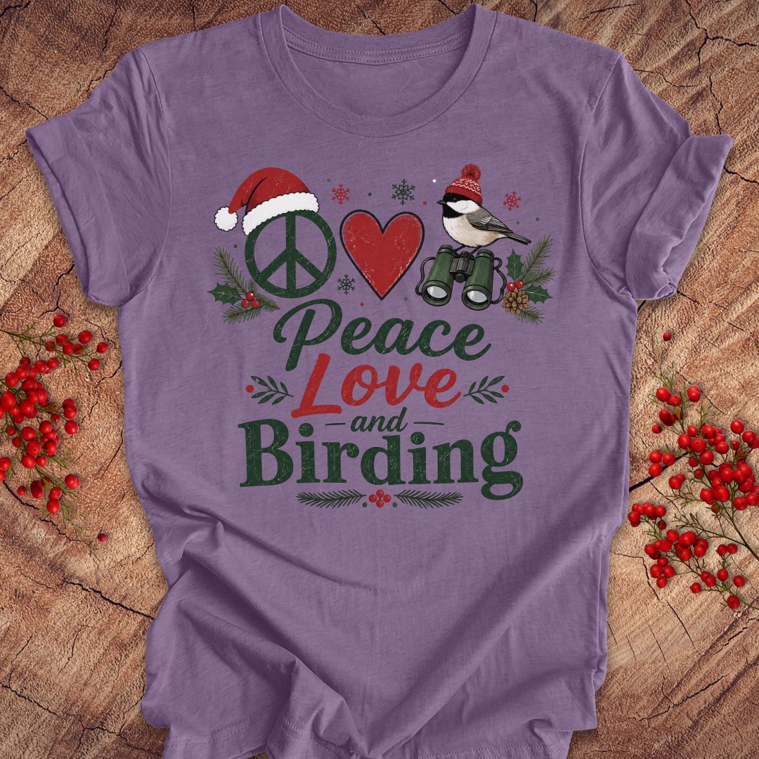 A purple short-sleeve t-shirt laid flat on a rustic wood background, decorated with red berries. The shirt features a graphic design with a peace sign wearing a Santa hat, a red heart, a bird with binoculars, and the words 'Peace Love and Birding' in green and red lettering.