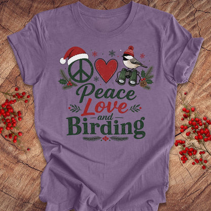 A purple short-sleeve t-shirt laid flat on a rustic wood background, decorated with red berries. The shirt features a graphic design with a peace sign wearing a Santa hat, a red heart, a bird with binoculars, and the words 'Peace Love and Birding' in green and red lettering.
