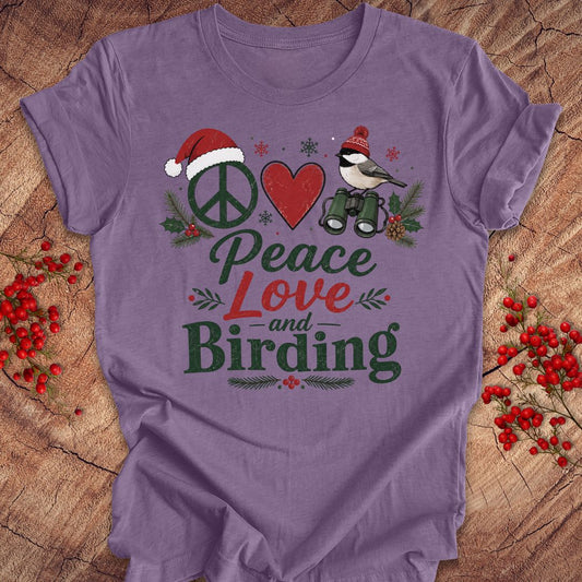 A purple short-sleeve t-shirt laid flat on a rustic wood background, decorated with red berries. The shirt features a graphic design with a peace sign wearing a Santa hat, a red heart, a bird with binoculars, and the words 'Peace Love and Birding' in green and red lettering.