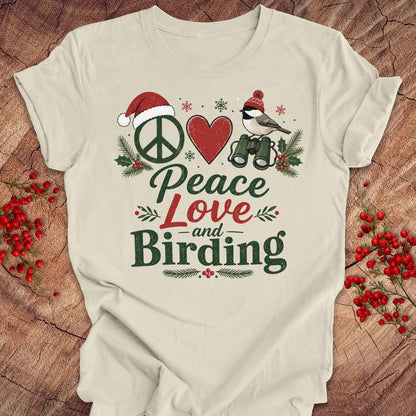A cream-colored short-sleeve t-shirt with a festive Christmas birding graphic that reads 'Peace Love and Birding'. The graphic features a peace sign in a Santa hat, a red heart, a chickadee with binoculars, and holiday greenery, laid flat on a rustic wood background with red berries.