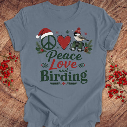 A slate blue t-shirt with a festive Christmas birding graphic on the front, laid flat on a rustic wood background decorated with red berries. The graphic features a peace sign with a Santa hat, a red heart, a chickadee on binoculars, and the text 'Peace Love and Birding'.