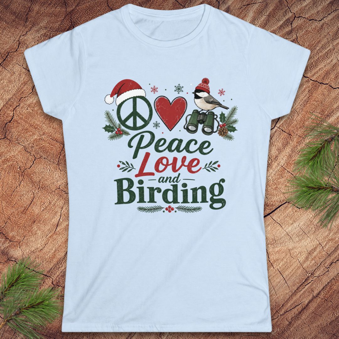 A light blue women's short-sleeve t-shirt with a festive Christmas birding graphic on the front, laid flat on a rustic wood background with pine branches in the corners. The graphic features a peace sign, a red heart, a bird with binoculars, and the words 'Peace Love and Birding'.