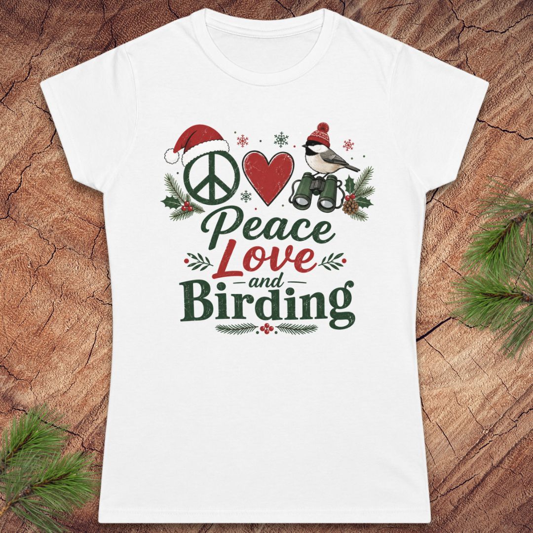 A white women's short-sleeve T-shirt laid flat on a rustic wood surface with pine branches around it. The shirt features a colorful front graphic reading 'Peace Love and Birding' with a peace sign, red heart, bird, Santa hat, and holiday greenery.