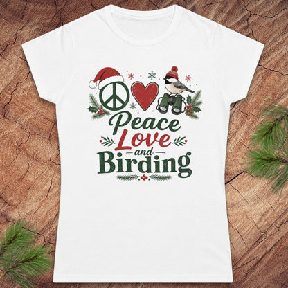 A white women's short-sleeve T-shirt laid flat on a rustic wood surface with pine branches around it. The shirt features a colorful front graphic reading 'Peace Love and Birding' with a peace sign, red heart, bird, Santa hat, and holiday greenery.