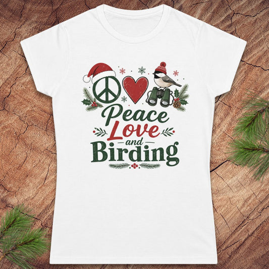 A white women's short-sleeve T-shirt laid flat on a rustic wood surface with pine branches around it. The shirt features a colorful front graphic reading 'Peace Love and Birding' with a peace sign, red heart, bird, Santa hat, and holiday greenery.