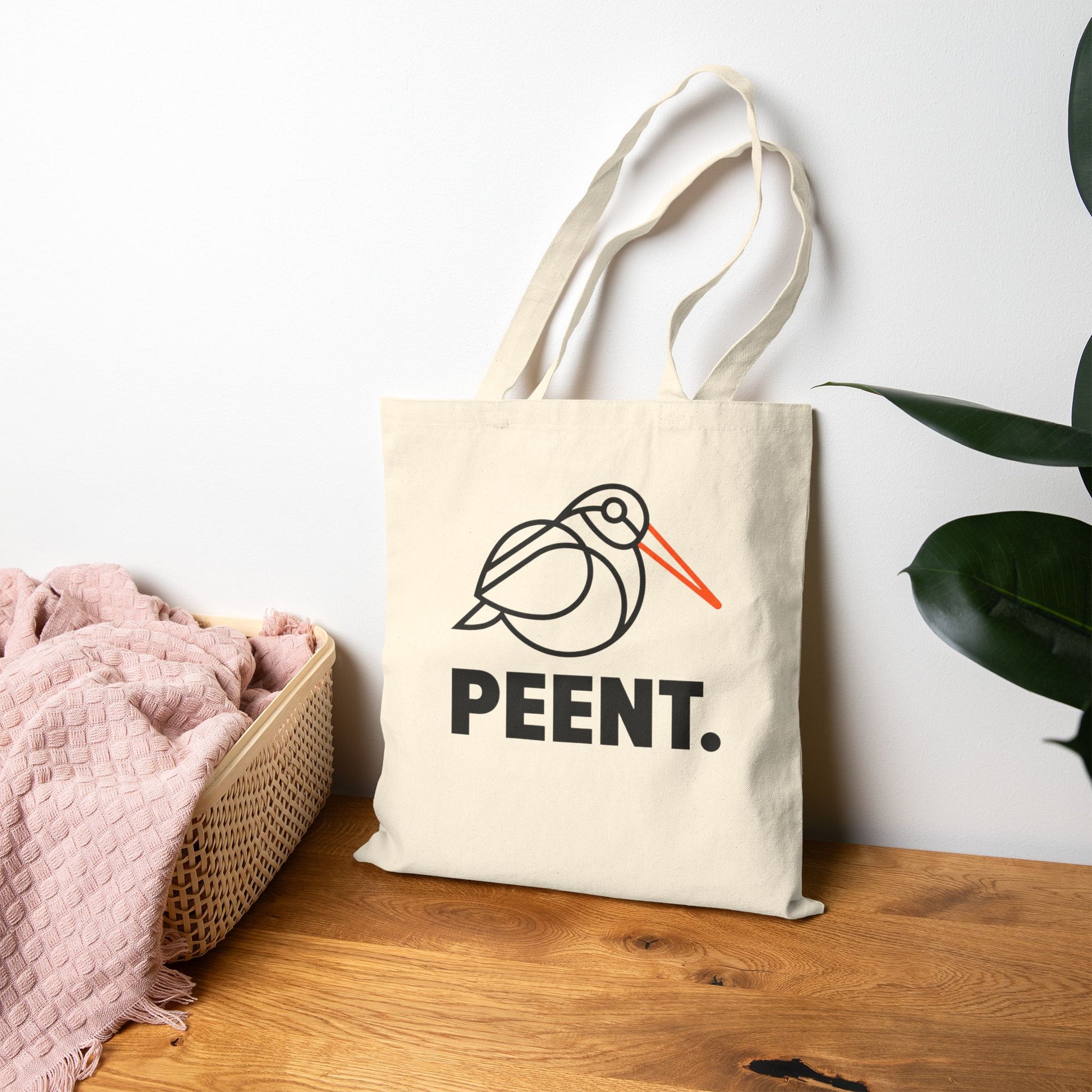 Tote bag with 'PEENT.' design on a wooden surface with a white wall and plant in the background