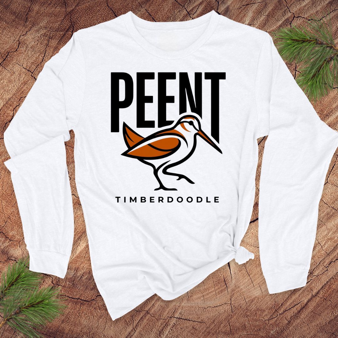 White long-sleeve shirt with 'PEENT' and 'TIMBERDOODLE' design on a wooden surface.