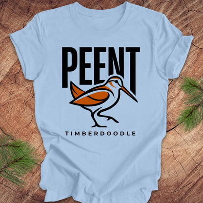 Light blue t-shirt with 'PEENT' and 'TIMBERDOODLE' woodcock design on a wooden surface.