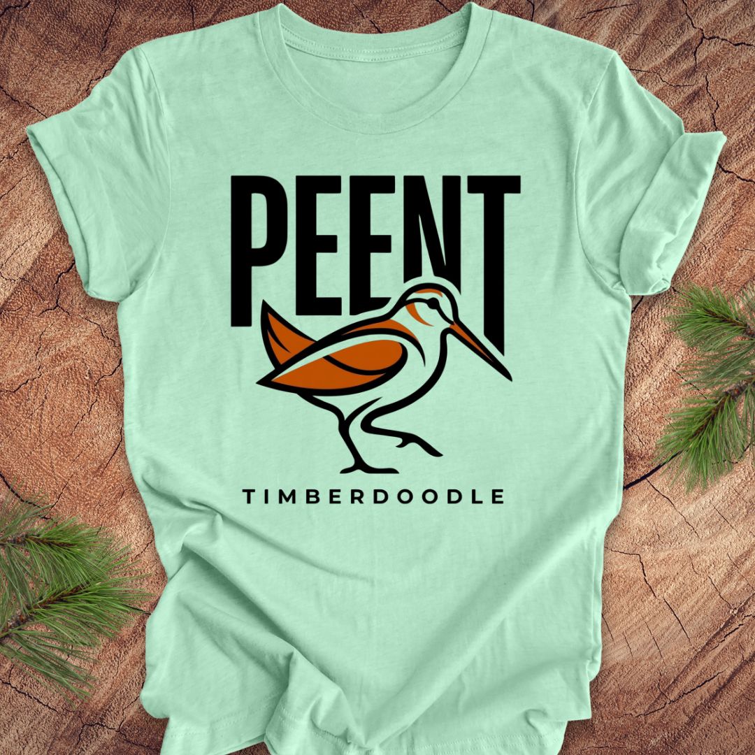Light green t-shirt with 'PEENT' and 'TIMBERDOODLE' woodcock design on a wooden surface.
