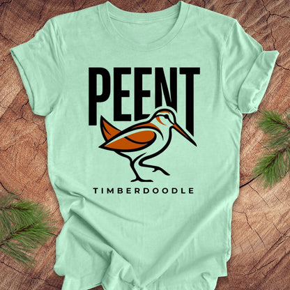Light green t-shirt with 'PEENT' and 'TIMBERDOODLE' woodcock design on a wooden surface.
