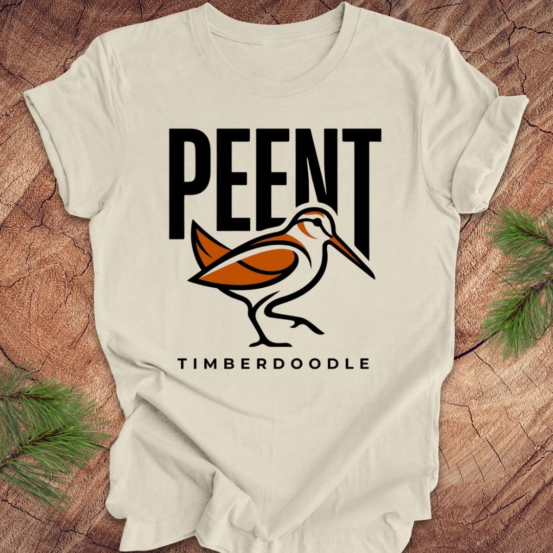 Beige t-shirt with 'PEENT' and 'TIMBERDOODLE' woodcock design on a wooden surface.
