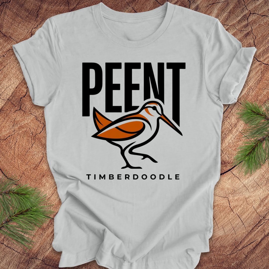 Silver t-shirt with 'PEENT' and 'TIMBERDOODLE' woodcock design on a wooden surface.