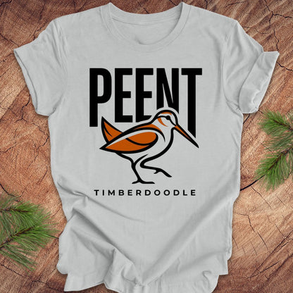 Silver t-shirt with 'PEENT' and 'TIMBERDOODLE' woodcock design on a wooden surface.