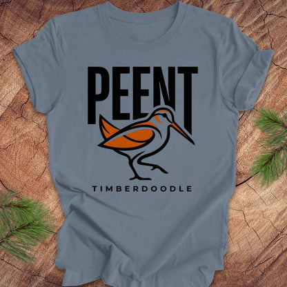 Steel blue t-shirt with 'PEENT' and 'TIMBERDOODLE' woodcock design on a wooden surface.