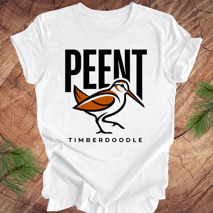 White t-shirt with 'PEENT' and 'TIMBERDOODLE' woodcock design on a wooden surface.