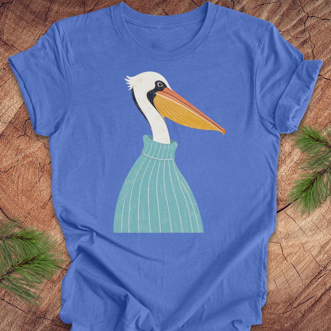 A royal blue short-sleeve t-shirt laid flat on a rustic wood surface, featuring a stylized graphic of a white pelican with a yellow-orange beak and a light green striped sweater. Pine branches are visible in the corners.