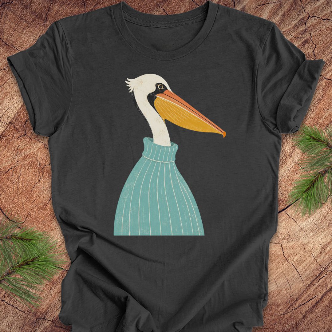 A dark charcoal gray t-shirt featuring a stylized graphic of a white pelican wearing a teal striped sweater, laid flat on a rustic wood background with pine branches in the corners.