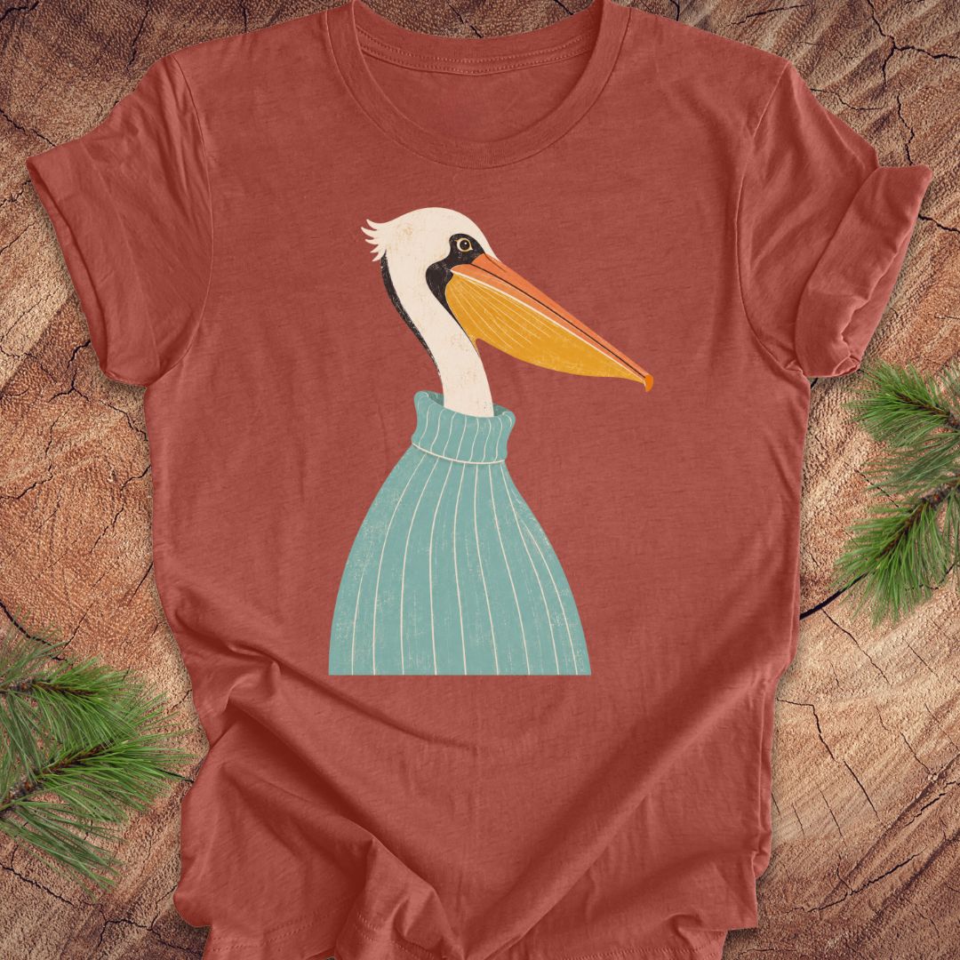 A heathered terracotta short-sleeve t-shirt featuring a stylized graphic of a white pelican with a yellow-orange beak and a teal striped sweater, laid flat on a rustic wood background with pine branches.