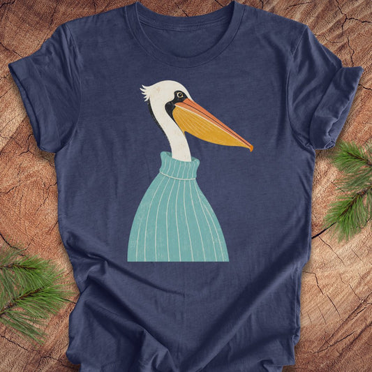A navy blue short-sleeve t-shirt laid flat on a rustic wood surface, featuring a stylized graphic of a white pelican with a yellow-orange beak and a teal striped sweater. Green pine branches are placed in the corners of the frame.