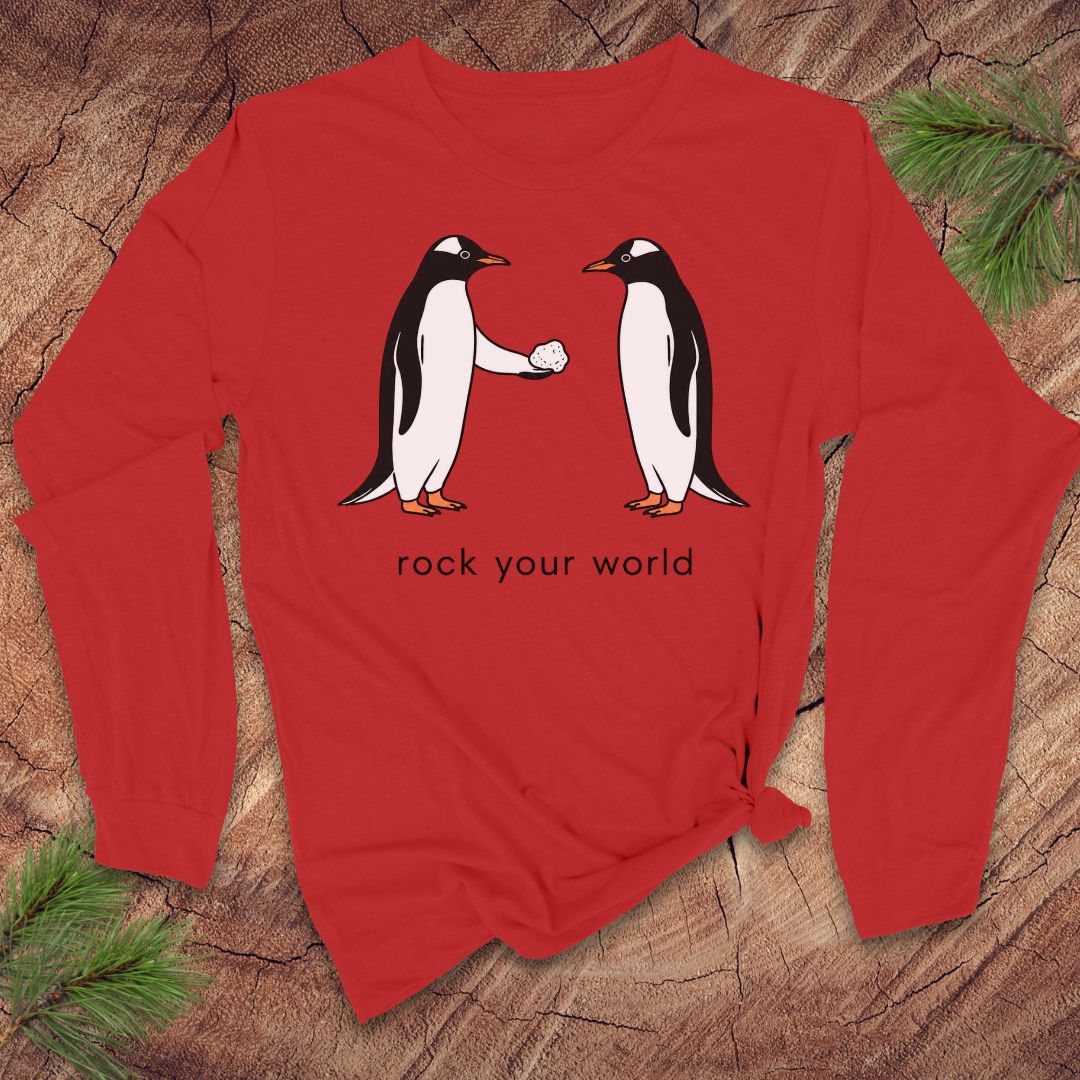 Red long-sleeve t-shirt with penguins and text 'rock your world' on a wood background.
