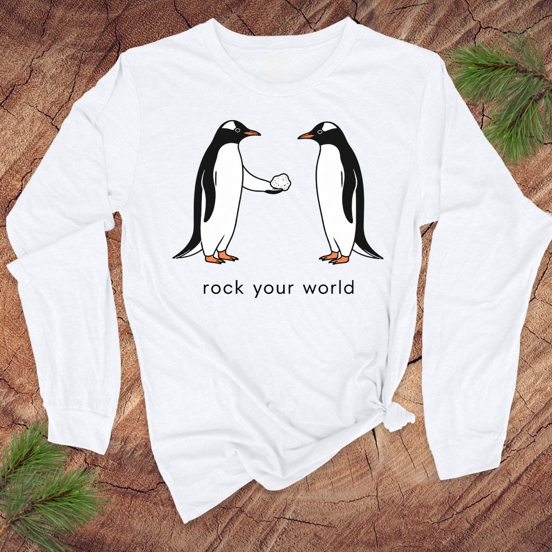White long-sleeve t-shirt with penguins and text 'rock your world' on a wood background.