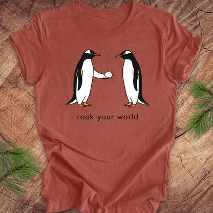 Rust t-shirt with a penguin handing another penguin a rock with text, 'rock your world' on a wood background.