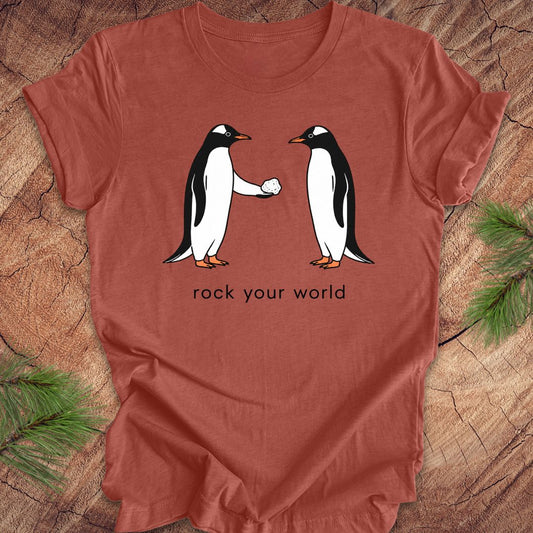 Rust t-shirt with a penguin handing another penguin a rock with text, 'rock your world' on a wood background.