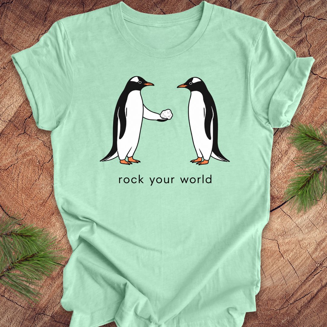 Mint green t-shirt with a penguin handing another penguin a rock with text, 'rock your world' on a wood background.
