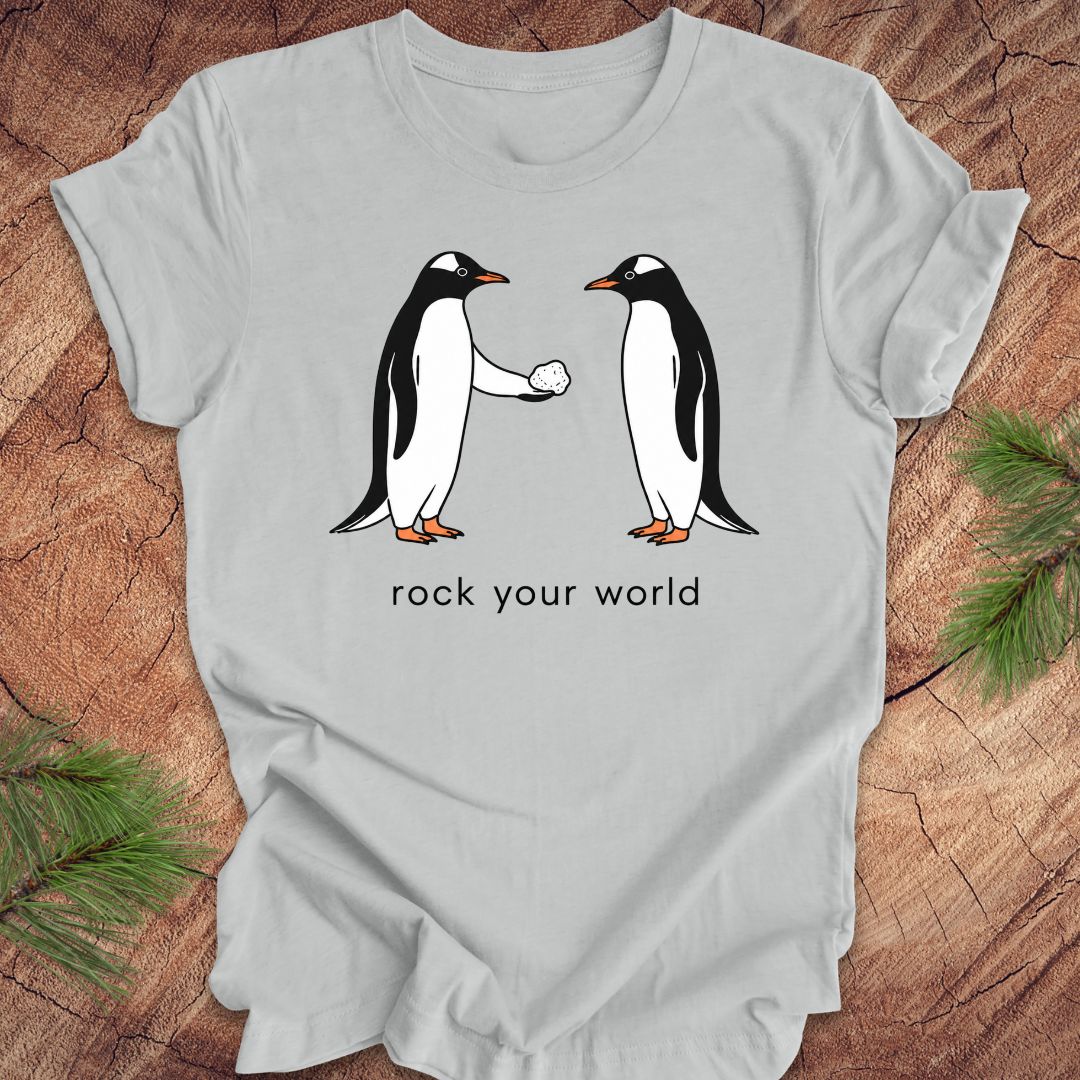 Gray t-shirt with a penguin handing another penguin a rock with text, 'rock your world' on a wood background.