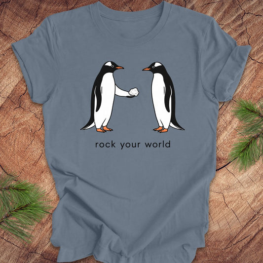Blue t-shirt with a penguin handing another penguin a rock with text, 'rock your world' on a wood background.