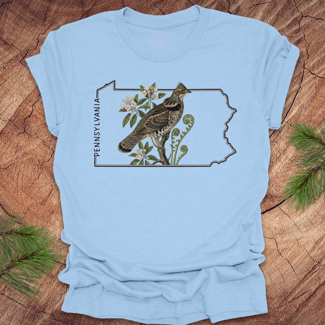 Baby blue t-shirt with a graphic of a ruffed grouse and plants on a Pennsylvania outline design, placed on a wooden surface.