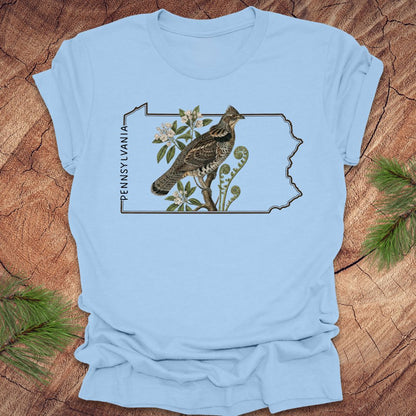 Baby blue t-shirt with a graphic of a ruffed grouse and plants on a Pennsylvania outline design, placed on a wooden surface.