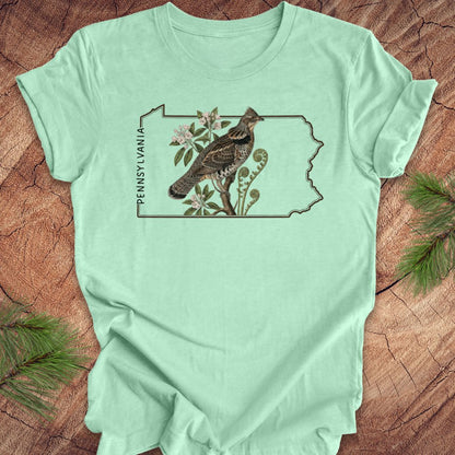 Mint green t-shirt with the state outline of Pennsylvania with ruffed grouse and floral design on a wooden surface