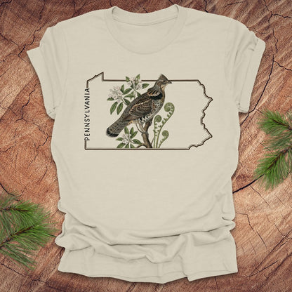 Natural t-shirt with a graphic of a ruffed grouse and plants on a Pennsylvania outline design, placed on a wooden surface.