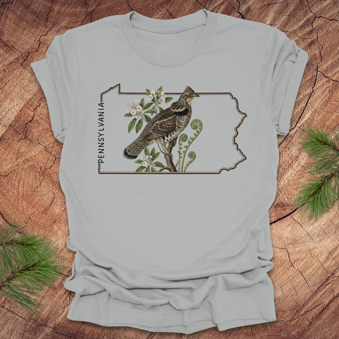 Silver t-shirt with a graphic of a ruffed grouse and plants on a Pennsylvania outline design, placed on a wooden surface.
