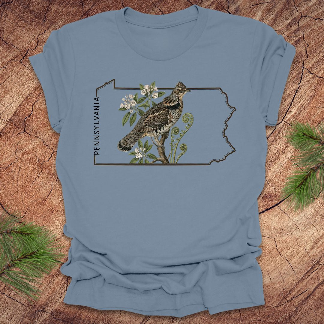 Steel blue t-shirt with a graphic of a ruffed grouse and plants on a Pennsylvania outline design, placed on a wooden surface.