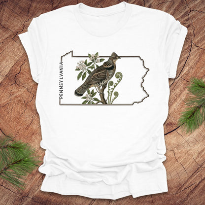 White t-shirt with a graphic of a ruffed grouse and plants on a Pennsylvania outline design, placed on a wooden surface.