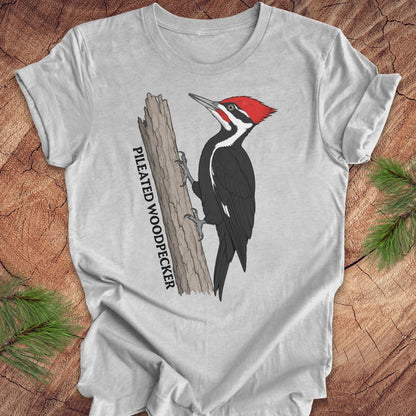 A light gray short-sleeve t-shirt laid flat on a rustic wood surface, featuring a graphic of a pileated woodpecker with a red crest and black-and-white body, plus vertical text reading 'PILEATED WOODPECKER.' Green pine branches are placed around the shirt as styling props.
