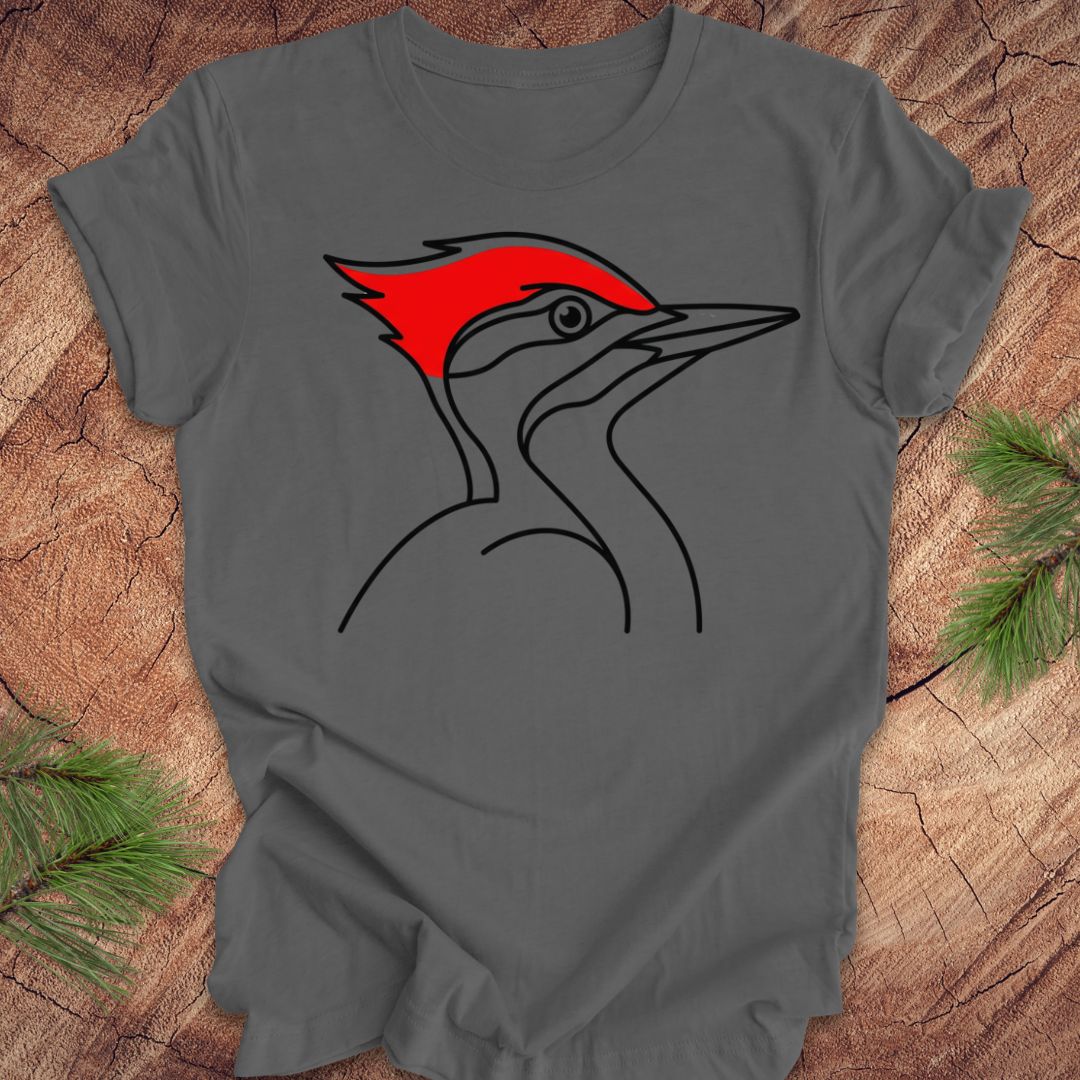 A dark gray short-sleeve t-shirt featuring a minimalist black line art graphic of a woodpecker with a bright red crest, laid flat on a rustic wood background with pine needles in the corners.